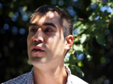 Picture of Cedar Sigo standing outdoors in front of a background of green leaves and sunlight. The person's face is blurred, and they are wearing a light-colored plaid shirt.